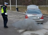 Policjant w umundurowaniu służbowym, w białej czapce stoi przy samochodzie osobowym, z którego rury wydechowej wydobywają się kłęby szarego dymu
