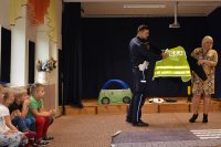 Policjant z profilaktykiem mówią dzieciom o bezpiecznej drodze, pokazują kamizelkę odblaskową