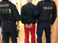 Policjanci w umundurowaniu ćwiczebnym prowadzą korytarzem mężczyznę w kajdankach
