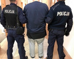 Dwóch policjantów w umundurowaniu służbowym doprowadza korytarzem zatrzymanego mężczyznę w jasnych spodniach i ciemnej bluzie