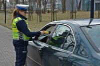 Policjanta w umundurowaniu służbowym, w białej czapce, sprawdza stan trzeźwości urządzeniem ALCOBLOW, kierującego samochodem osobowym koloru ciemnozielonego
