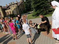 Na podwórku policjant w umundurowaniu ćwiczebnym (czarnym) wspólnie z psem służbowym prowadzi spotkanie z grupą dzieci. Z prawej strony siostra zakonna w białym habicie.