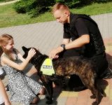 Policjant w umundurowaniu ćwiczebnym (czarnym) przykucnięty przy psie służbowym, którego głaszcze dziewczynka o długich włosach w letniej sukience