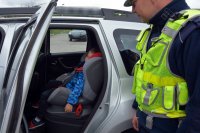 Policjant w umundurowaniu służbowym stoi przy dziecku, siedzącym w samochodzie, w foteliku