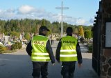 Policjanci w umundurowaniu służbowym, w kamizelkach odblaskowych, patrolują cmentarz