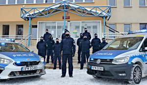 Grupa policjantów w umundurowaniu ćwiczebnym z psami służbowymi na tle wejścia do budynku Komendy Miejskiej Policji w Suwałkach, stoją pomiędzy dwoma radiowozami