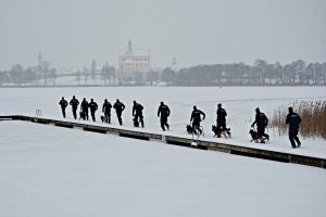 Grupa policjantów w umundurowaniu ćwiczebnym z psami służbowymi biegnie po terenie ośnieżonego Wigierskiego Parku Narodowego, w tle klasztor