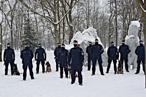 Grupa policjantów w umundurowaniu ćwiczebnym z psami służbowymi stoi w parku, na tle białego niedźwiedzia