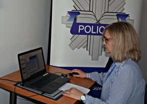 Policyjny profilaktyk, kobieta w okularach siedzi przy laptopie, w tle plakat z gwiazda policyjną