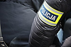 Ręka policjantka w czarnej kurtce z opaską, z napisem POLICJA, siedzący w samochodzie mężczyzna w czarnej kurtce