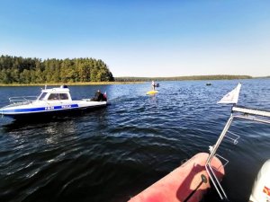 Policyjna łódź służbowa na jeziorze, w tle łódź WOPR i przewrócona żaglówka, na której jest dwóch mężczyzn