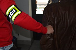 Policjant w czerwonej bluzie z opaską na ręku z napisem POLICJA doprowadza zatrzymanego mężczyznę, z rękoma w kajadankach, ubranego w brązową kurtkę