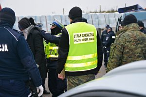 Policjanci, strażnicy graniczni wykonują czynności z zatrzymanymi przy czerwonym busie
