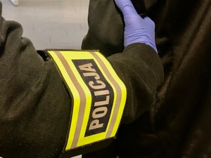 Ręka policjanta z opaską odblaskową, na której jest napis POLICJA, doprowadzającego zatrzymanego