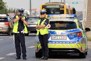 Policjanci stoją przy radiowozie, mierzą prędkość jadących pojazdów