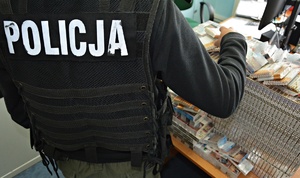 Policjant po cywilnemu w czarnej kamizelce z napisem POLICJA stoi obok biurka, na którym są nielegalne papierosy.