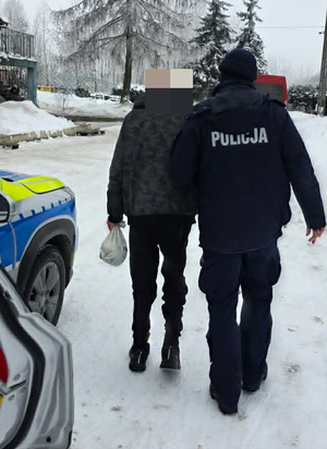 Policjant pomaga iść mężczyźnie w tle budynki oraz pojazdy